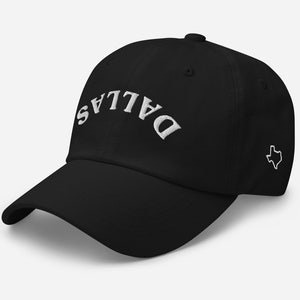 May include: Black baseball cap with the word "DALLAS" embroidered in white across the front. A white outline of the state of Texas is embroidered on the side. The cap has a curved brim and an adjustable strap.