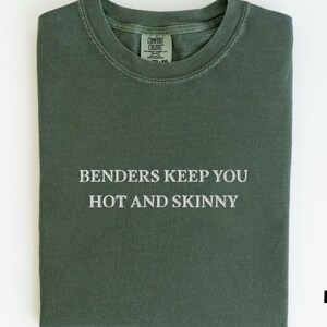 Puede incluir: Una camiseta verde musgo doblada con el texto "Benders Keep You Hot and Skinny" en blanco. La camiseta tiene cuello redondo y una etiqueta en la parte superior de la espalda. La palabra "Moss" está impresa en la esquina inferior derecha.