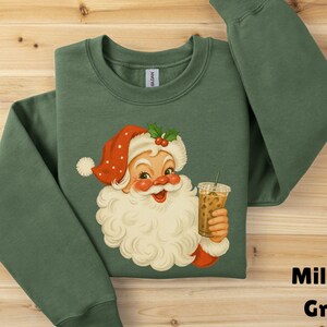 Puede incluir: Una sudadera de cuello redondo color verde militar con una ilustración gráfica de Santa Claus que lleva un gorro rojo con lunares blancos y sostiene un vaso de plástico transparente lleno de café helado con una pajita, centrado en el pecho.