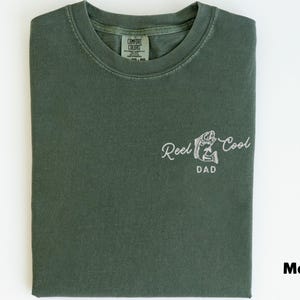 May include: A moss green t-shirt with the words "Reel Cool Dad" in white script, along with a fish graphic. The shirt is folded neatly and has a tag in the upper center.