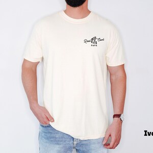 Op de afbeelding: Ivoor kleurig t-shirt met de tekst "Reel Cool Papa" in zwart script. Het t-shirt heeft een ronde hals. De persoon die het t-shirt draagt, draagt een blauwe spijkerbroek en een horloge.