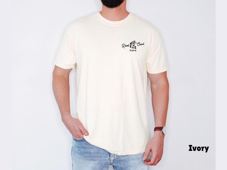 Op de afbeelding: Ivoor kleurig t-shirt met de tekst "Reel Cool Papa" in zwart script. Het t-shirt heeft een ronde hals. De persoon die het t-shirt draagt, draagt een blauwe spijkerbroek en een horloge.