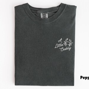 May include: A folded, dark gray t-shirt with the phrase "A Little Crabby" and a crab graphic embroidered in white on the front. The shirt is made of a soft material and has a classic crew neck.