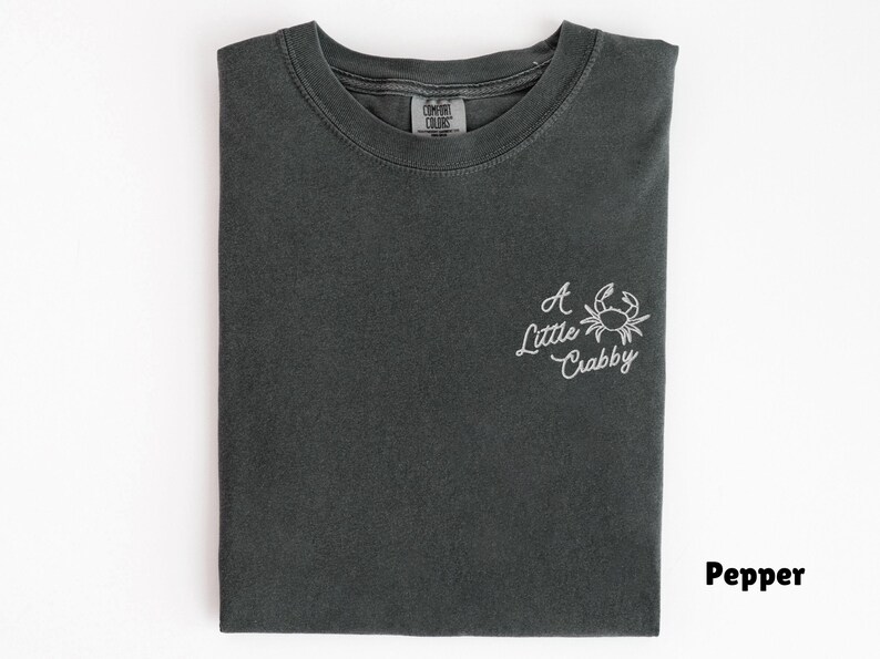 May include: A folded, dark gray t-shirt with the phrase "A Little Crabby" and a crab graphic embroidered in white on the front. The shirt is made of a soft material and has a classic crew neck.
