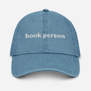 May include: A blue denim baseball cap with white embroidered text that reads "book person".