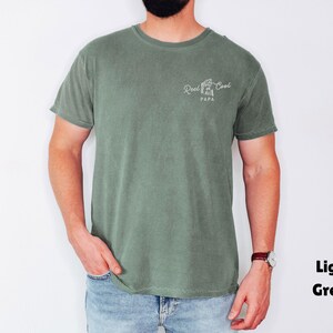 Op de afbeelding: Lichtgroen t-shirt met de tekst "Reel Cool Papa" in witte letters. Het shirt is effen van kleur en heeft een ronde hals. De persoon draagt een blauwe spijkerbroek en een horloge.