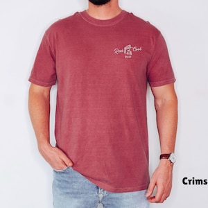 Pu&ograve; includere: T-shirt color cremisi con la scritta "Reel Cool Dad" in corsivo bianco. La maglietta a maniche corte e girocollo &egrave; indossata con jeans blu. Il modello indossa un orologio con cinturino in pelle marrone.