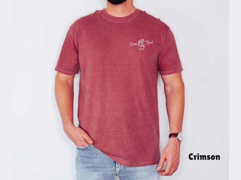 Pu&ograve; includere: T-shirt color cremisi con la scritta "Reel Cool Dad" in corsivo bianco. La maglietta a maniche corte e girocollo &egrave; indossata con jeans blu. Il modello indossa un orologio con cinturino in pelle marrone.
