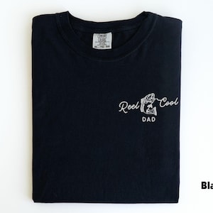 Pu&ograve; includere: T-shirt nero con la scritta "Reel Cool Dad" in bianco. La maglietta ha una piccola grafica di un pesce. La maglietta &egrave; piegata e ha un'etichetta in alto al centro.