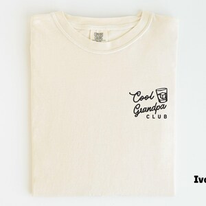 May include: Ivory-colored t-shirt with the text "Cool Grandpa Club" in black script and a graphic of a drink. The shirt is folded and displayed on a white background. The shirt is made of a soft material.