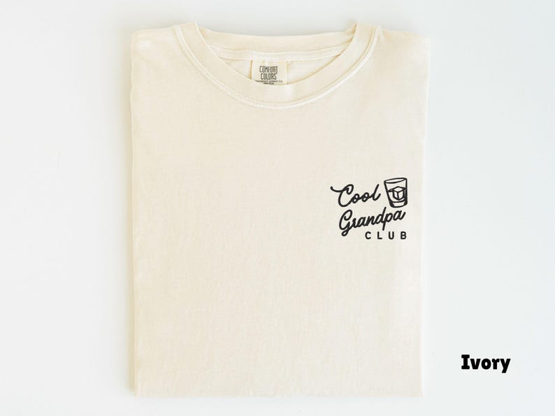 May include: Ivory-colored t-shirt with the text "Cool Grandpa Club" in black script and a graphic of a drink. The shirt is folded and displayed on a white background. The shirt is made of a soft material.