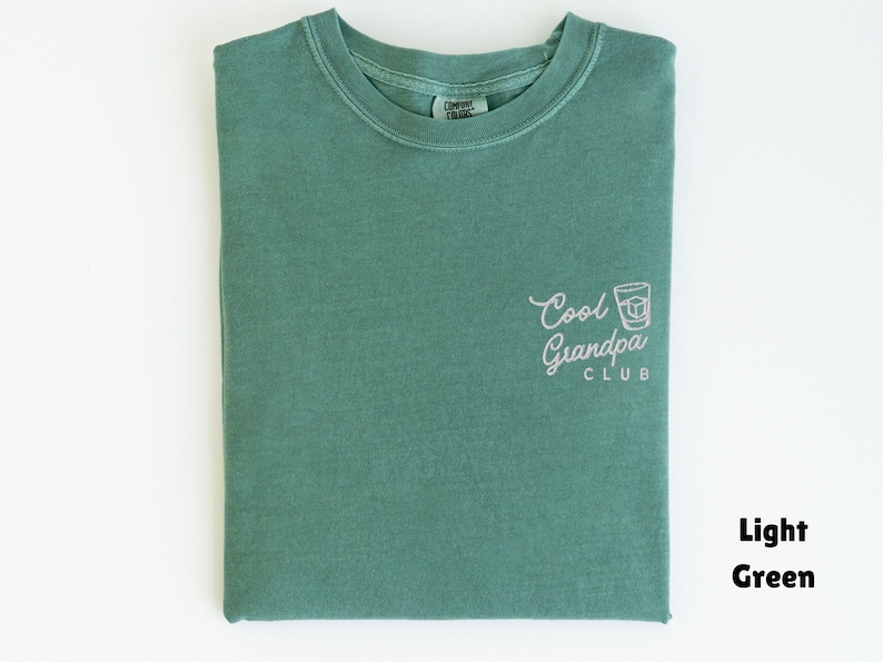 May include: Light green t-shirt with the text "Cool Grandpa Club" in white script and a graphic of a whiskey glass. The shirt is folded and has a crew neck. The shirt is made of a soft material.