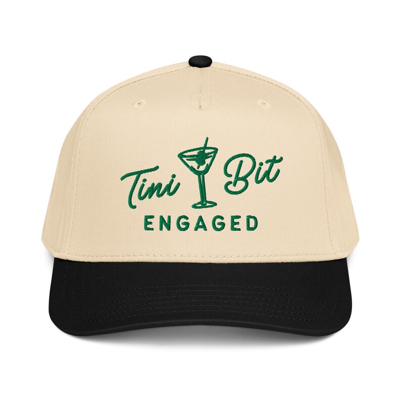 Puede incluir: Gorra de b&eacute;isbol con corona color crema y visera negra. La gorra presenta las palabras "Tini Bit Engaged" en verde, junto con un gr&aacute;fico de copa de martini. La gorra tiene un bot&oacute;n negro en la parte superior.