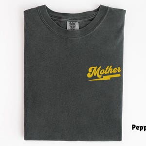 May include: A folded, dark gray t-shirt with the word "Mother" in yellow script and a yellow lightning bolt graphic. The shirt is made of a soft material and has a classic crew neck.