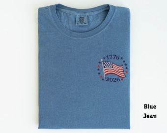 May include: Blue jean colored t-shirt with a small embroidered American flag design. The flag is surrounded by stars and the numbers "1776" and "2026". The shirt is folded neatly on a white surface.