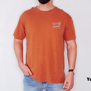 May include: A burnt orange t-shirt with the text "Cool Grandpa Club" printed on the upper left chest. The shirt is short-sleeved and has a crew neck. The model is wearing blue jeans and a watch.