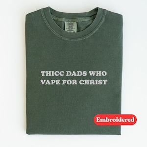 Puede incluir: Una camiseta verde oscuro con el texto bordado "THICC DADS WHO VAPE FOR CHRIST". La camiseta est&aacute; doblada cuidadosamente sobre una superficie blanca. Una pegatina ovalada roja que dice "Embroidered" est&aacute; en la parte inferior derecha.