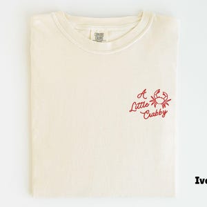May include: Ivory-colored t-shirt with a red crab graphic and the text "A Little Crabby." The shirt is folded and has a crew neck. The Comfort Colors label is visible at the neckline.