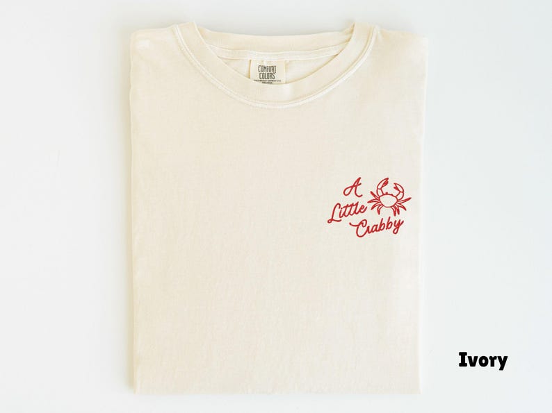 May include: Ivory-colored t-shirt with a red crab graphic and the text "A Little Crabby." The shirt is folded and has a crew neck. The Comfort Colors label is visible at the neckline.