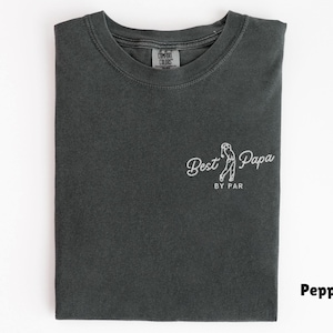 Op de afbeelding: Een gevouwen, peperkleurig t-shirt met de witte tekst "Best Papa by Par" en een golf-thema illustratie. Het t-shirt heeft een ronde hals en een label met "Comfort Colors" is zichtbaar. Het ontwerp staat op de voorkant.
