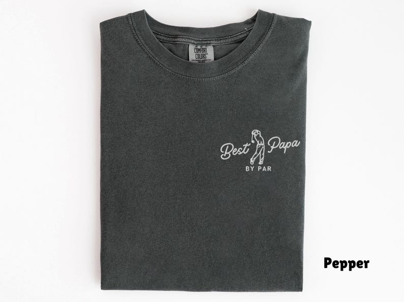 Op de afbeelding: Een gevouwen, peperkleurig t-shirt met de witte tekst "Best Papa by Par" en een golf-thema illustratie. Het t-shirt heeft een ronde hals en een label met "Comfort Colors" is zichtbaar. Het ontwerp staat op de voorkant.