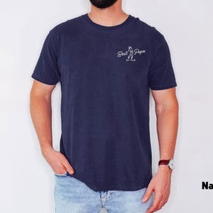 Op de afbeelding: Marineblauw T-shirt met de tekst "Best Papa" en een golfclubafbeelding. Het shirt is effen van kleur en heeft een ronde hals. Het model draagt een blauwe spijkerbroek en een horloge.