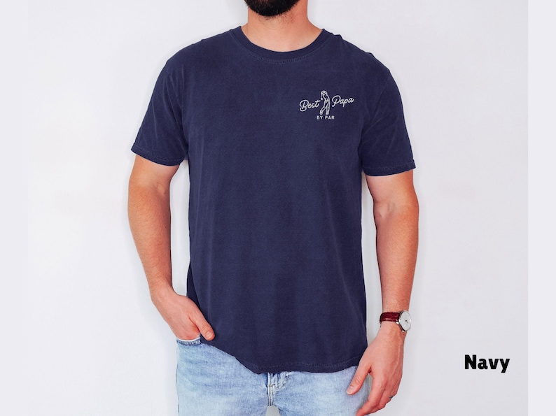 Op de afbeelding: Marineblauw T-shirt met de tekst "Best Papa" en een golfclubafbeelding. Het shirt is effen van kleur en heeft een ronde hals. Het model draagt een blauwe spijkerbroek en een horloge.