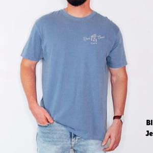 Op de afbeelding: Een blauw T-shirt met korte mouwen met de tekst "Reel Cool Papa" in witte letters. Het shirt wordt gecombineerd met een blauwe jeans. Het model draagt een horloge met een bruine leren band.