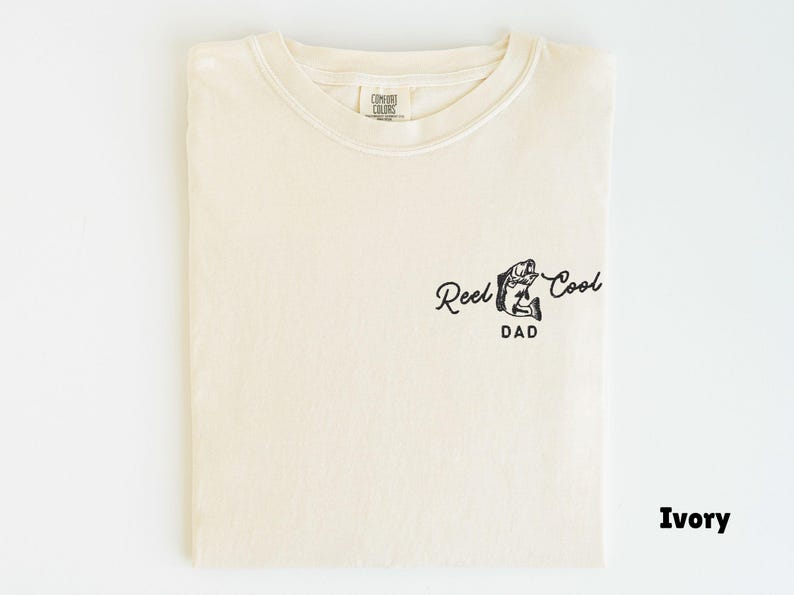Pu&ograve; includere: T-shirt color avorio con la scritta "Reel Cool Dad" e una grafica di un pesce. La maglietta &egrave; piegata e mostra il disegno sul davanti. Il tessuto sembra morbido e confortevole.