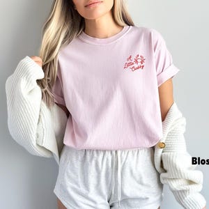 May include: A light pink t-shirt with the text "A Little Crabby" and a crab graphic in red. The shirt is paired with gray shorts and a white cardigan. The word "Blossom" is written in black.