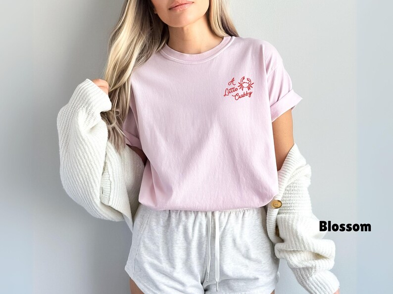 May include: A light pink t-shirt with the text "A Little Crabby" and a crab graphic in red. The shirt is paired with gray shorts and a white cardigan. The word "Blossom" is written in black.