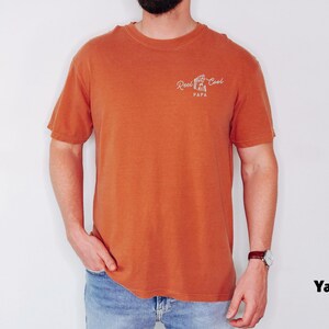 Op de afbeelding: Oranje T-shirt met korte mouwen met de tekst "Reel Cool Papa" in witte letters. Het shirt is effen van kleur en is gemaakt van een zacht materiaal. Het model draagt een blauwe spijkerbroek en een horloge.