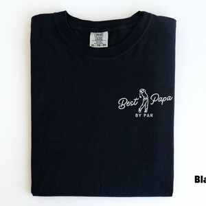 Op de afbeelding: Zwart T-shirt met de tekst "Best Papa by Par" in witte letters, met een afbeelding van een golfer. Het shirt is netjes opgevouwen en heeft een label in het midden. Het woord "Black" staat rechtsonder.