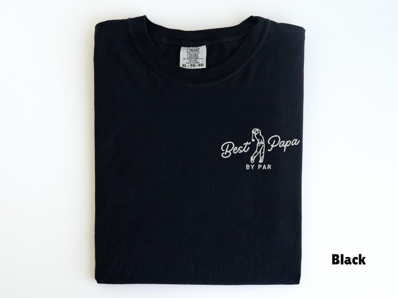 Op de afbeelding: Zwart T-shirt met de tekst "Best Papa by Par" in witte letters, met een afbeelding van een golfer. Het shirt is netjes opgevouwen en heeft een label in het midden. Het woord "Black" staat rechtsonder.