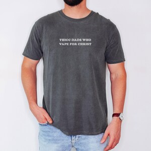 Puede incluir: Camiseta gris oscuro con el texto "THICC DADS WHO VAPE FOR CHRIST" en blanco. La camiseta es de manga corta y cuello redondo. La persona lleva vaqueros azules y un reloj con correa de cuero marr&oacute;n.