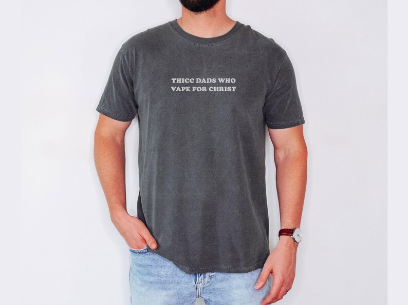 Puede incluir: Camiseta gris oscuro con el texto "THICC DADS WHO VAPE FOR CHRIST" en blanco. La camiseta es de manga corta y cuello redondo. La persona lleva vaqueros azules y un reloj con correa de cuero marr&oacute;n.
