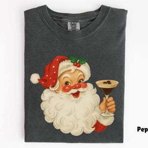 May include: Dark gray t-shirt featuring a vintage illustration of Santa Claus holding a martini. Santa is wearing a red hat with white polka dots and a white beard. The martini has coffee beans. The shirt is a Comfort Colors brand.