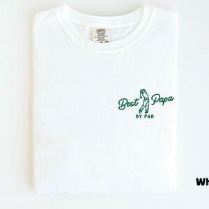 Op de afbeelding: Wit T-shirt met de tekst "Best Papa by Par" in groen en een golfswing-afbeelding. Het shirt is opgevouwen en toont het ontwerp op de voorkant. Het shirt is gemaakt van een zacht materiaal.
