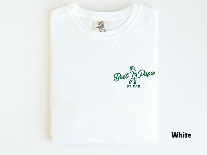 Op de afbeelding: Wit T-shirt met de tekst "Best Papa by Par" in groen en een golfswing-afbeelding. Het shirt is opgevouwen en toont het ontwerp op de voorkant. Het shirt is gemaakt van een zacht materiaal.