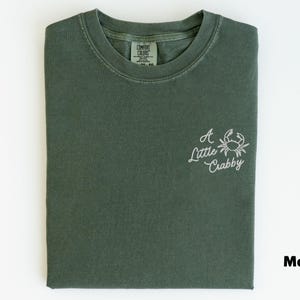 May include: Moss green t-shirt with the phrase "A Little Crabby" and a crab graphic embroidered in white. The shirt is folded neatly and has a Comfort Colors tag.