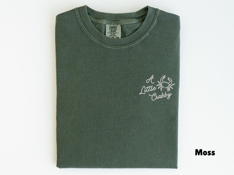May include: Moss green t-shirt with the phrase "A Little Crabby" and a crab graphic embroidered in white. The shirt is folded neatly and has a Comfort Colors tag.