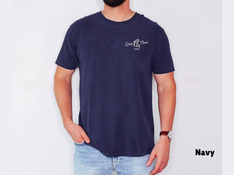 Pu&ograve; includere: T-shirt blu navy con la scritta "Reel Cool Dad" in corsivo bianco. La maglietta ha girocollo e maniche corte. Il modello indossa jeans blu e un orologio con cinturino in pelle marrone.