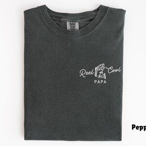 Op de afbeelding: Pepperkleurig T-shirt met een ronde hals. Het shirt toont de tekst "Reel Cool Papa" in een cursief lettertype, samen met een visafbeelding. Het shirt is opgevouwen en heeft een label bij de halslijn.