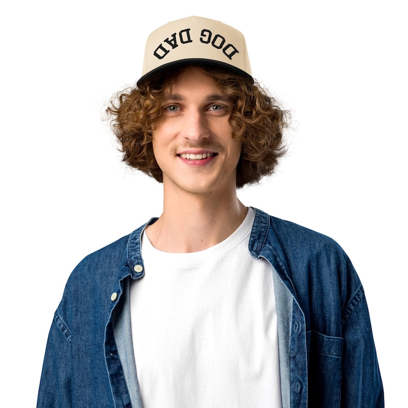 May include: A beige flat-billed cap with the words "DOG DAD" in black block letters. The cap has a black brim. The person in the image is wearing a white t-shirt and a denim jacket.