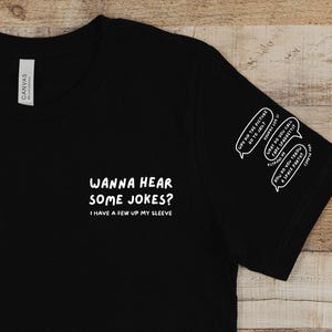 May include: Black t-shirt with white text: "WANNA HEAR SOME JOKES? I HAVE A FEW UP MY SLEEVE." The sleeve has three speech bubbles with jokes. The shirt is made of a soft material.