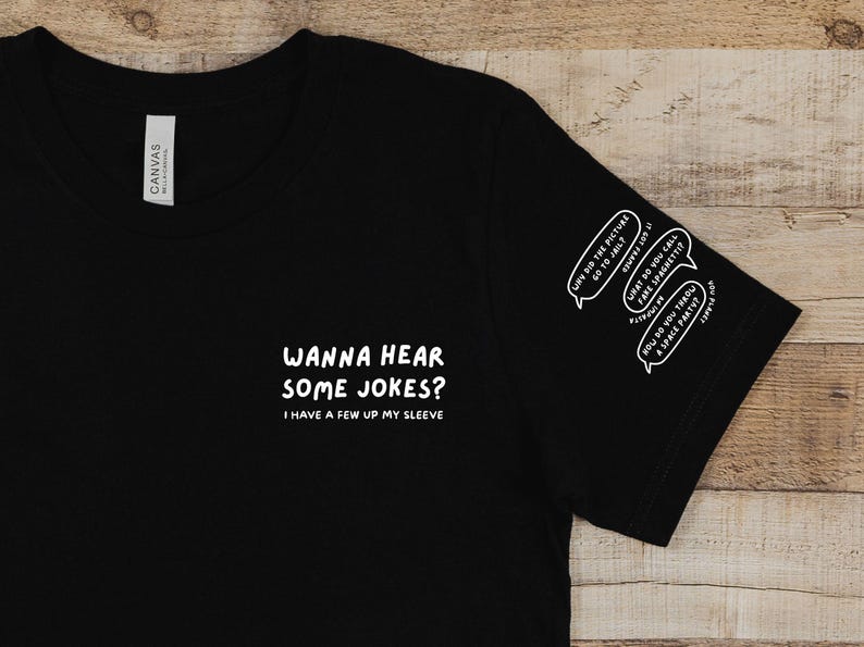 May include: Black t-shirt with white text: "WANNA HEAR SOME JOKES? I HAVE A FEW UP MY SLEEVE." The sleeve has three speech bubbles with jokes. The shirt is made of a soft material.