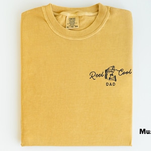Pu&ograve; includere: T-shirt color senape con la scritta "Reel Cool Dad" e una grafica di pesce. La maglietta &egrave; piegata e ha un'etichetta nella parte superiore centrale. Il colore &egrave; indicato come "Mustard" in basso a destra.