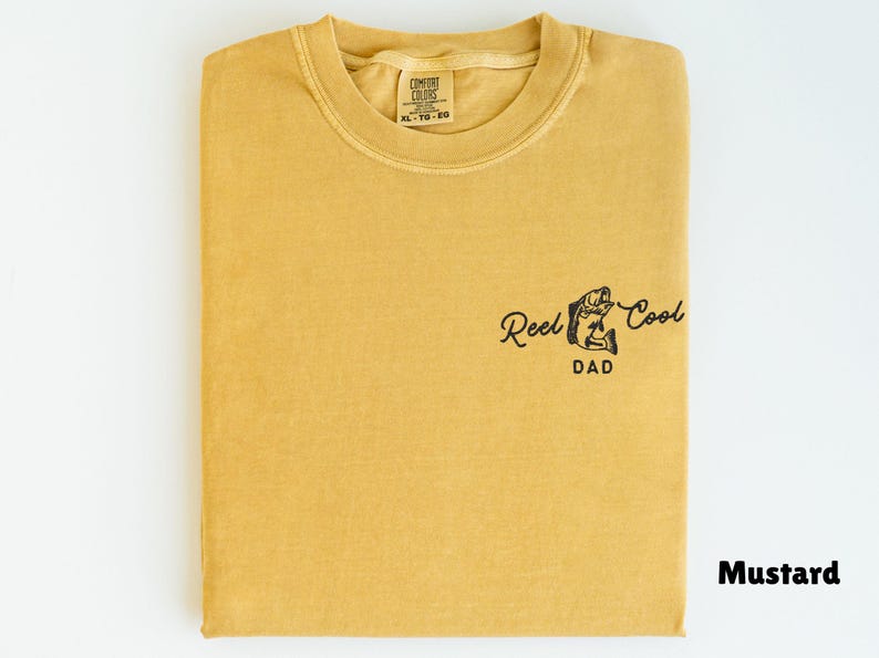 Pu&ograve; includere: T-shirt color senape con la scritta "Reel Cool Dad" e una grafica di pesce. La maglietta &egrave; piegata e ha un'etichetta nella parte superiore centrale. Il colore &egrave; indicato come "Mustard" in basso a destra.