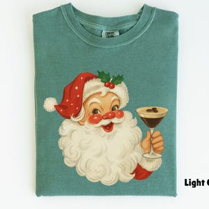 May include: Light green t-shirt featuring a vintage illustration of Santa Claus holding a cocktail. Santa is wearing a red hat with white trim and a white beard. The cocktail has coffee beans on top. The text "Light Green" is at the bottom right.