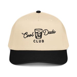 May include: A beige baseball cap with a black brim. The cap features the embroidered text "Cool Dads CLUB" in black script and block letters, along with a graphic of a cocktail glass with an ice cube.
